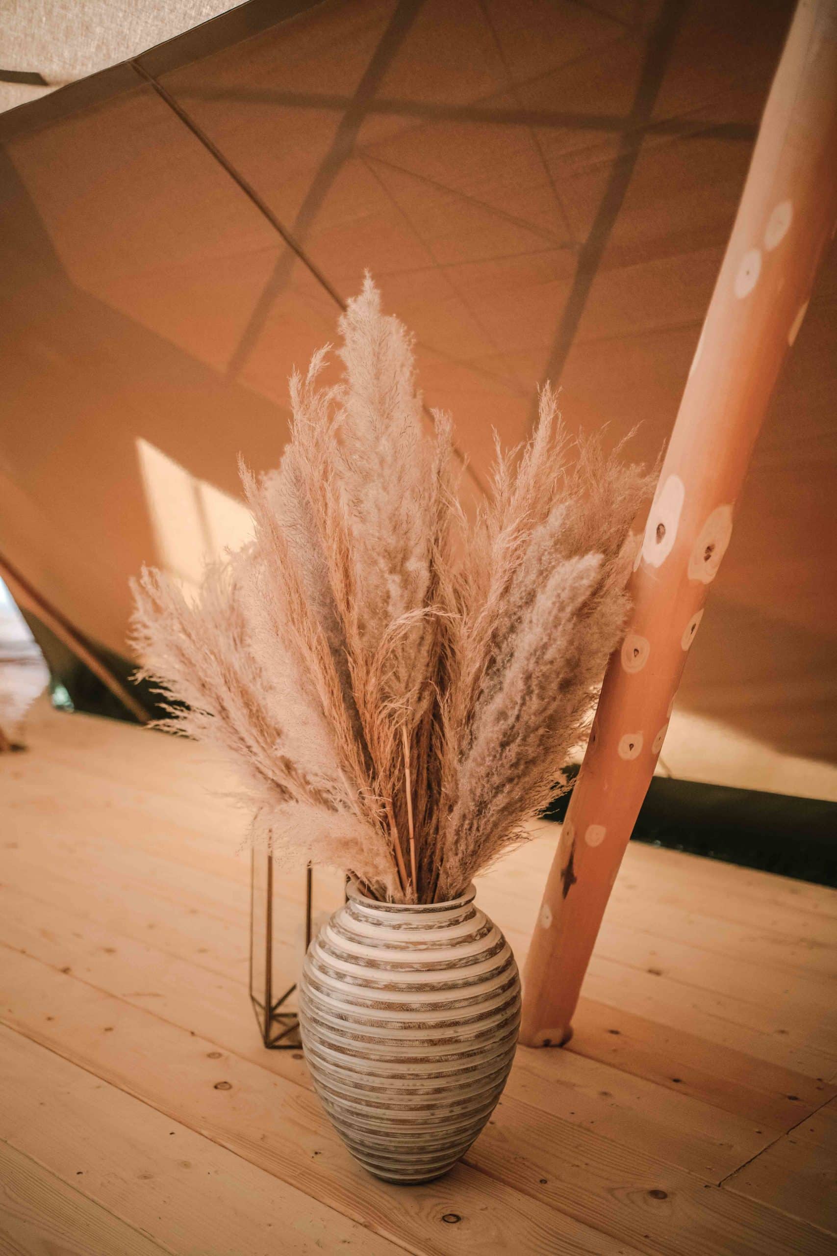 Wonderland Wedding Venues | A large, elongated vase with horizontal, textured stripes, sitting on a wooden floor, is filled with dried pampas grass. The background includes part of a tent structure and a decorated wooden pole. Warm, natural light fills the scene, creating a cozy ambiance.
