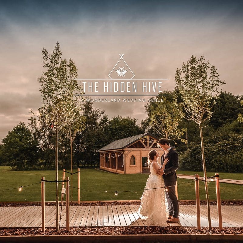 Wonderland Wedding Venues | A bride and groom stand on a wooden pathway, surrounded by lush greenery. They gaze at each other lovingly under a dramatic sky filled with clouds. The pathway is adorned with lanterns and leads to a wooden cabin in the background. Text overlays read "The Hidden Hive.