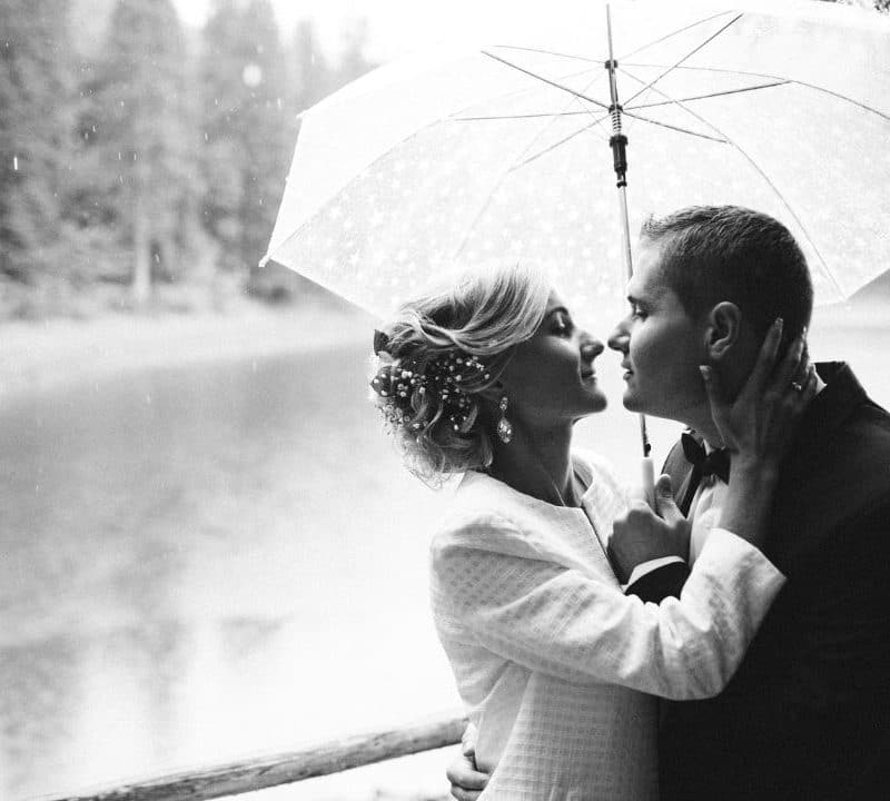 Wonderland Wedding Venues | Black and white image of a couple under a transparent umbrella by a lake. The woman, in a dress and coat, and the man, in a suit, are about to kiss. Trees and water are visible in the blurry background, creating a romantic and intimate atmosphere.