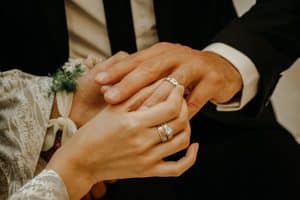 Wonderland Wedding Venues | Close-up of two people holding hands, focusing on their intertwined fingers. Both are wearing wedding bands, symbolizing marriage or engagement. One hand is adorned with intricate jewelry and a floral wrist accessory. Both individuals are dressed in formal attire.