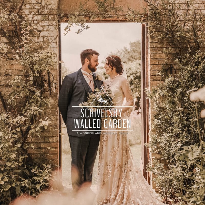 Wonderland Wedding Venues | A bride and groom stand facing each other, holding hands in an arched doorway surrounded by lush greenery at Scrivelsby Walled Garden. They are both smiling and dressed in wedding attire, with the bride in a white gown and the groom in a dark suit.