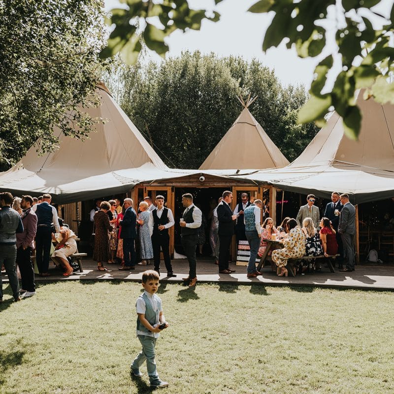 Wonderland Wedding Venues | A group of people are gathered outside a large, open-sided tent adorned with a trio of peaked roofs on a sunny day at a popular wedding venue in Northamptonshire. They are dressed in semi-formal attire, socializing and enjoying the event. A young boy stands in the foreground on a grassy area, holding a small item.