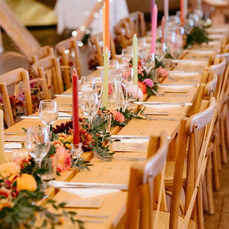 Wonderland Wedding Venues | A long wooden table is elegantly set for a meal, adorned with colorful candles and vibrant floral arrangements, perfect for showcasing the work of wedding suppliers in Lincolnshire. Wooden chairs line both sides, and twinkling string lights are visible in the background, creating a warm and festive atmosphere.
