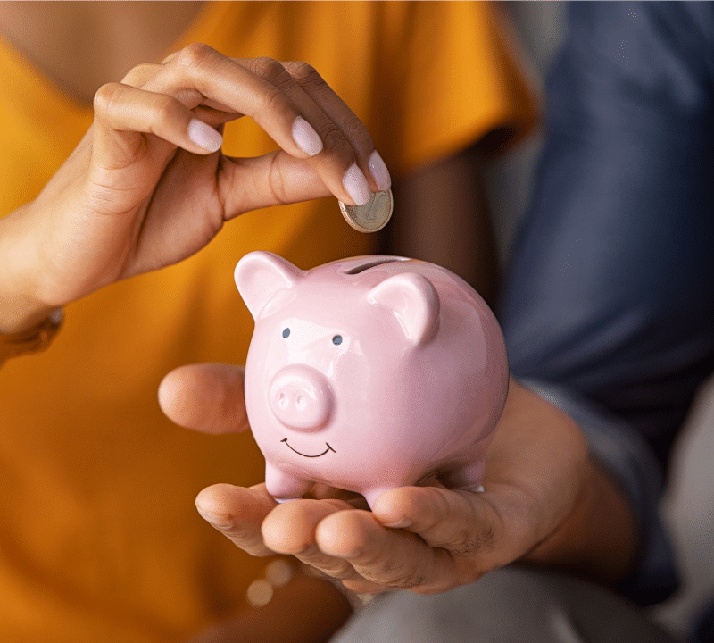 Wonderland Wedding Venues | Close-up of a person wearing a yellow shirt placing a coin into a pink piggy bank held by another person. The piggy bank is smiling, and the scene suggests saving money.
